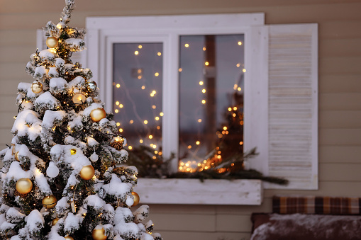 A snow-covered Christmas tree decorated with toys stands on the porch of a country white wooden cottage. The shuttered window is framed with garlands.