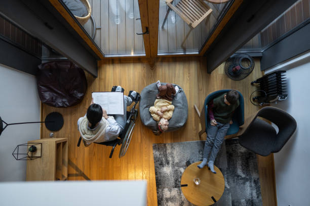 High angle view of disabled young mother reading book in wheelchair while her kids, son and little daughter relaxing at home