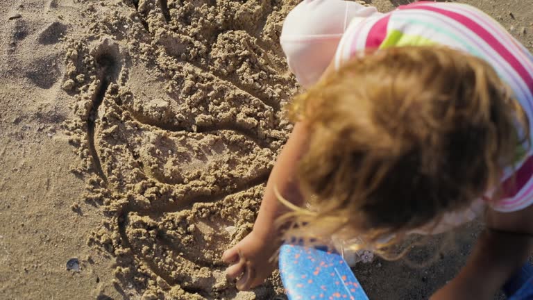 Little girl draws something on sand by the beach. Top view