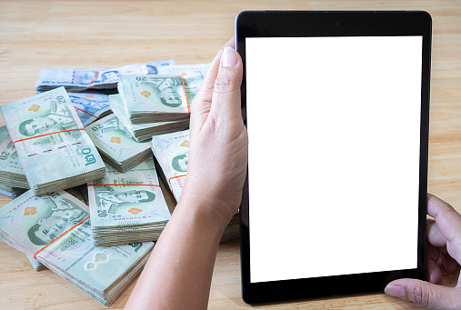 Close up of woman hands with white screen smartphone, tablet and stack of Thai banknotes. business, finance, saving, technology and people concept