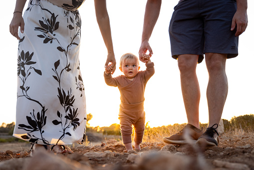 A baby girl is holding hands with her parents while learning how to walk. Back lit scene at sunset. Low angle.