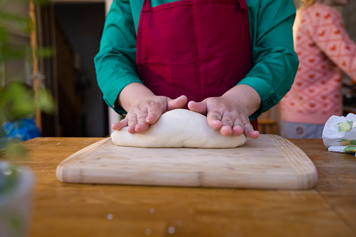 Close-up of a dumpling dough being moulded on a cutting board in a kitchen by an unrecognisable person wearing a green blouse and a red apron.
