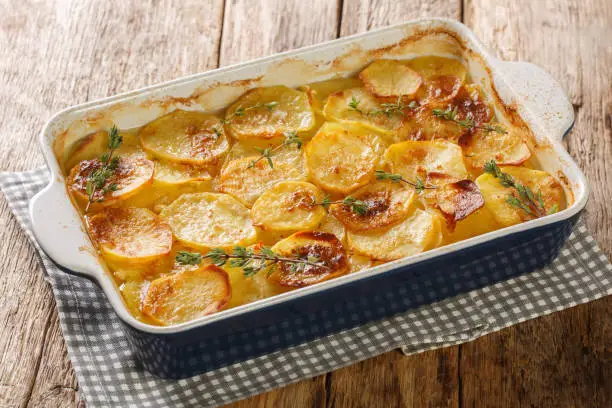 Scalloped potatoes, potato casserole with the addition of herbs, onion and garlic in a ceramic baking dish closeup on the table. Horizontal