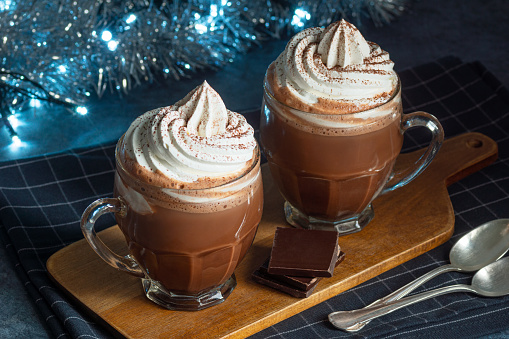 Two glass mugs of hot chocolate topped with whipped cream and cocoa powder. The glasses are on a wooden cutting board with pieces of a chocolate bar and festive holiday (Christmas or Hanukkah) decorations in the background.