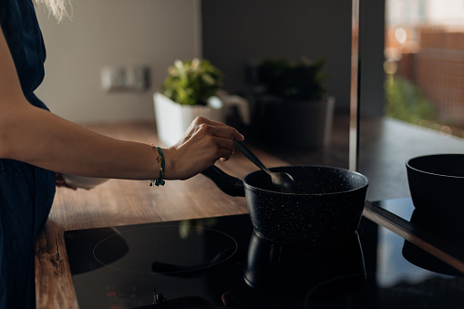 Close up of cropped unrecognizable woman hands cooking food in black stone frying pan on induction cooker, stove with ceramic top in modern kitchen. Chef preparing tasty diet food at home, new recipe
