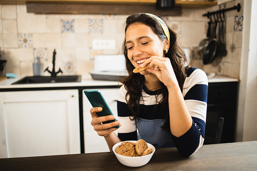 Portrait of a beautiful young woman snacking cookies and using smartphone at her kitchen