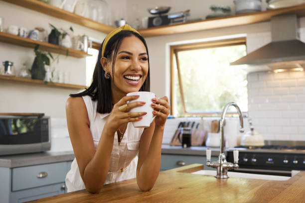 szczęśliwa kobieta, kawa lub herbata w domowej kuchni i relaks z uśmiechem rano w domu. spokojny młody człowiek, uśmiechnięty w spokoju i napój herbaty, aby obudzić się na początek dnia z pozytywnymi radosnymi myślami - serene people zdjęcia i obrazy z banku zdjęć