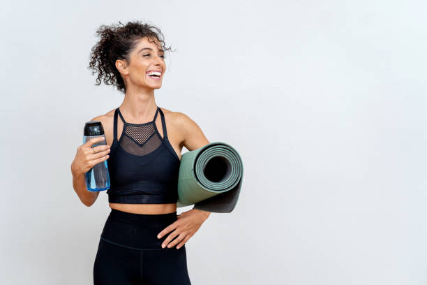 mujer sonriendo frente a la pared blanca con colchoneta y botella de agua - fitness apparel fotografías e imágenes de stock