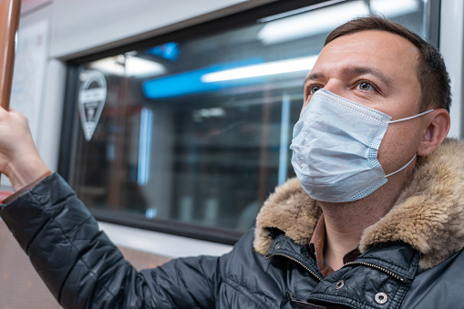 Portrait of male passenger wearing medical face mask in public transportation. Millennial man looking away at window, thinking, going to home by train during coronavirus pandemic. New normal