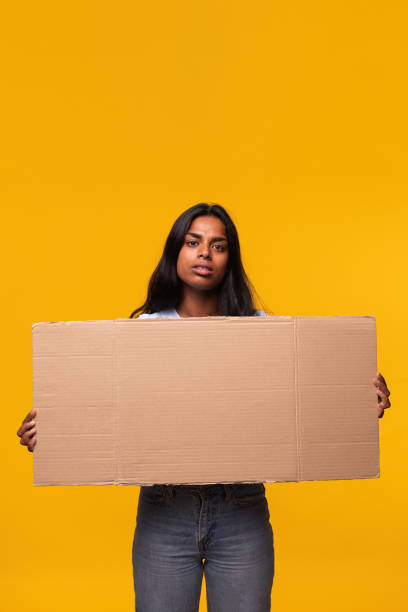 donna indiana con espressione seria che guarda la telecamera che tiene uno striscione di cartone isolato su sfondo giallo. - cardboard sign foto e immagini stock