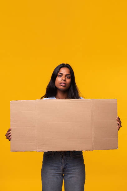 индийская женщина с серьезным выражением лица смотрит на камеру, держащую картонный баннер, изолированный на желтом фоне. - cardboard sign стоковые фото и изображения