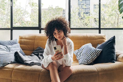 Young african american woman with curly hair sitting on a sofa in bright loft apartment living room looking cellphone with interest. Home concept. Technology concept.