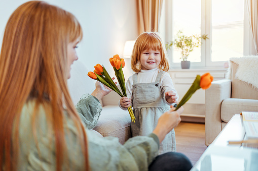 Portrait of a smiling mother and daughter embracing and holding a bouquet of fresh tulips at home. Child congratulates mother and gives a bouquet of flowers to tulips.