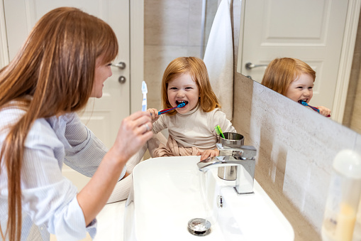 Shot of a mother and her little daughter brushing their teeth in the bathroom. Photo of a cute little girl and her mother teaching her how to brush teeth in the bathroom of their apartment; learning good personal hygiene habits at a young age.