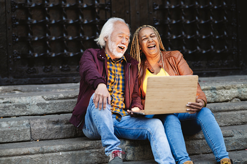 Multiracial senior couple laughing with a video in the laptop sitting in a stairs outdoors. Happy mature asian man and african american woman