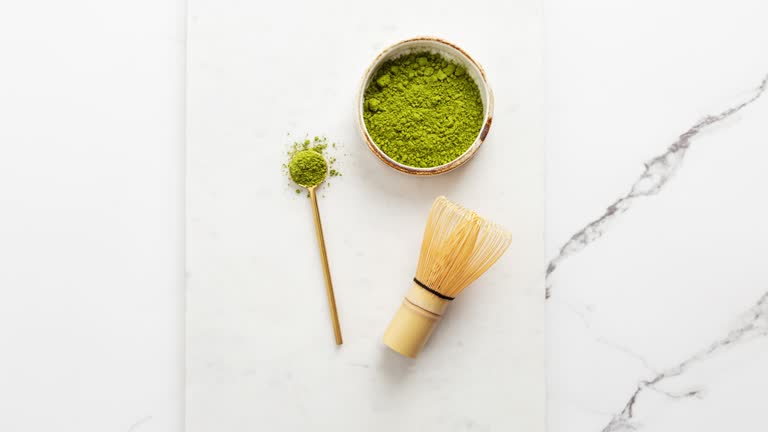 Matcha tea powder with spoon and bamboo whisk on white marble background