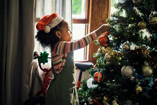 Little girl, Christmas and decorating tree for the festive season or holiday celebration at home. African American child hanging decor or lights for December or new year preparation at the house