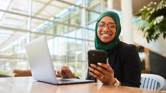 Young muslim businesswoman wearing a hijab smiling at a text while working on a laptop in an office lobby