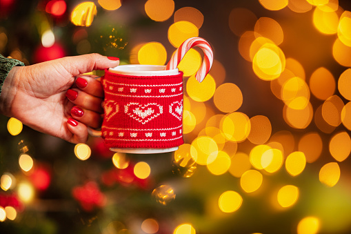 Close up view of woman's hand holding a red mug in front of defocused Christmas lights at background.