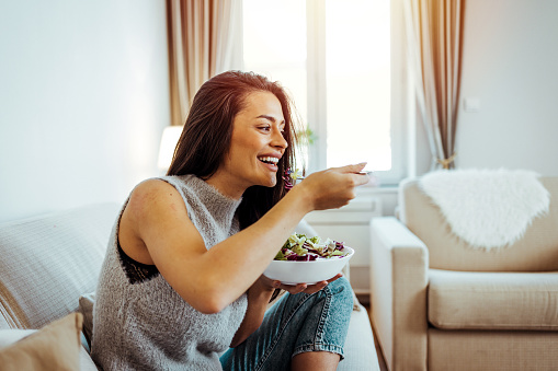 Healthy young woman dieting and eating a salad at home. Attractive woman with fresh green salad at home. A beautiful young woman is eating a salad in her living room, sitting on a sofa