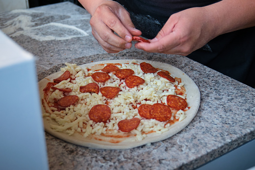 Close up of male hands adding pepperoni to a raw pizza in a restaurant kitchen. Pizzeria.