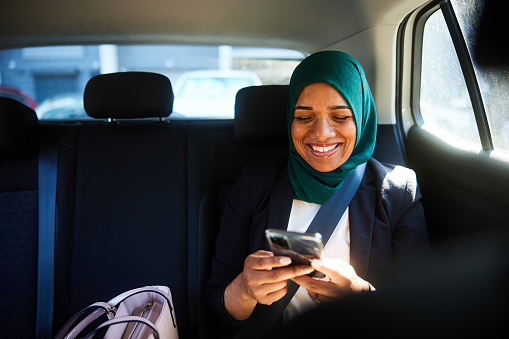 Young muslim businesswoman wearing a hijab smiling at a text on her phone while riding in the back seat of a taxi