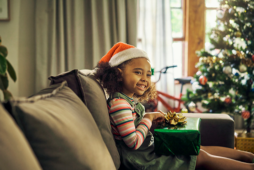 Christmas, child and gift or present on sofa while excited, happy and grateful in lounge with hat, tree and smile for holiday celebration. Black girl kid celebrate with gift box surprise on couch