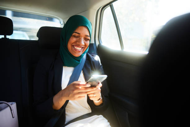 смеющаяся мусульманская бизнесвумен переписывается на свой телефон в задней части такси - mobile phone seat belt text messaging smiling стоковые фото и изображения