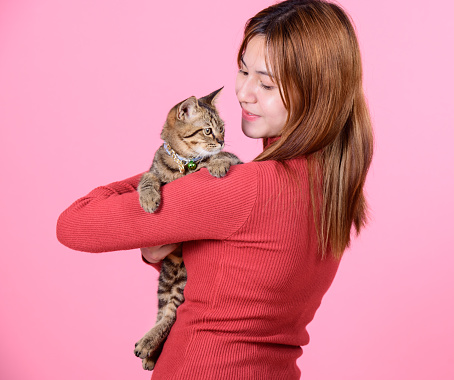 A young asian pretty cute woman in a red sweater holds a cat in her hands like a baby isolated on a bright color pink background. Asia girl with a kitten. Fiendship of pet and owner.