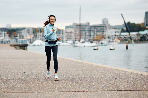 Happy Asian female runner with headphones jogging along a quay. Copy space.