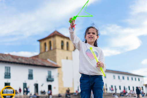 Latina girl with an average age of 7 years dressed casually stands in the historic square of Villa de Leyva boyaca playing with a bubbler to make giant bubbles while running and having fun in the breeze