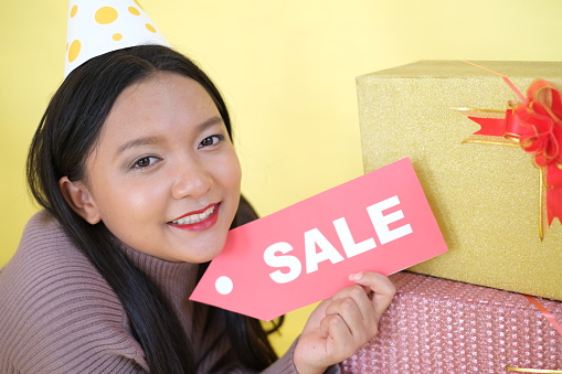 Happy young girl wear party hat hold sign and gift boxs on yellow background.