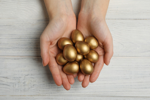 Woman holding shiny golden eggs at white wooden table, top view