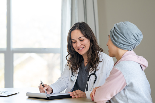 A female physician sits at a desk in her office and smiles while reviewing test results and sharing good news with an unrecognizable female patient recovering from cancer.