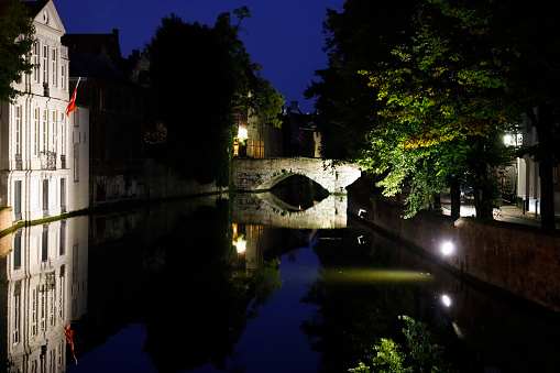 Bruges, Belgium - September 14, 2022: On a dark night in the old town you can see the illuminated stone bridge over the canal. Illuminated buildings and trees are also visible here