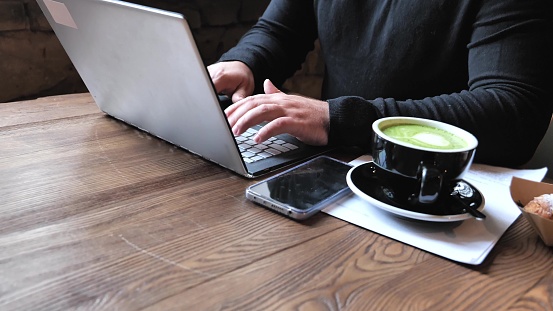 Male freelancer working with laptop computer, businessman work, typing on notebook while sitting at wooden table in modern coffee cafe with cup of coffee. Modern business, distance job, selfemployer. Selective focus