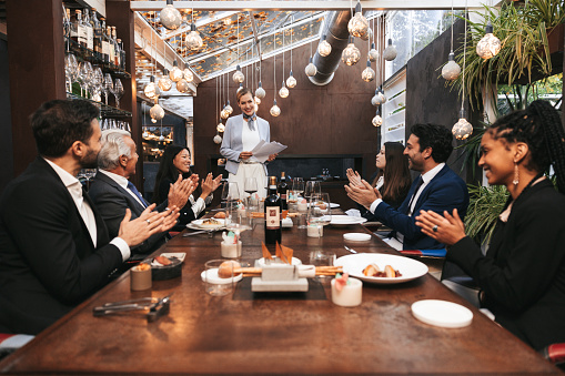 Business meeting in a luxury restaurant. Group of multi ethnic and diverse age range people attending to a corporate briefing sitting at a table after lunch.