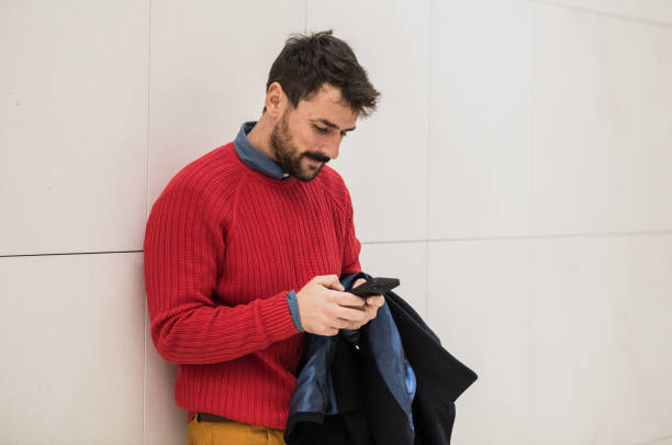 Young man leaning on a wall, while using his smart phone. Handsome young man in warm Winter clothes, using his mobile phone.