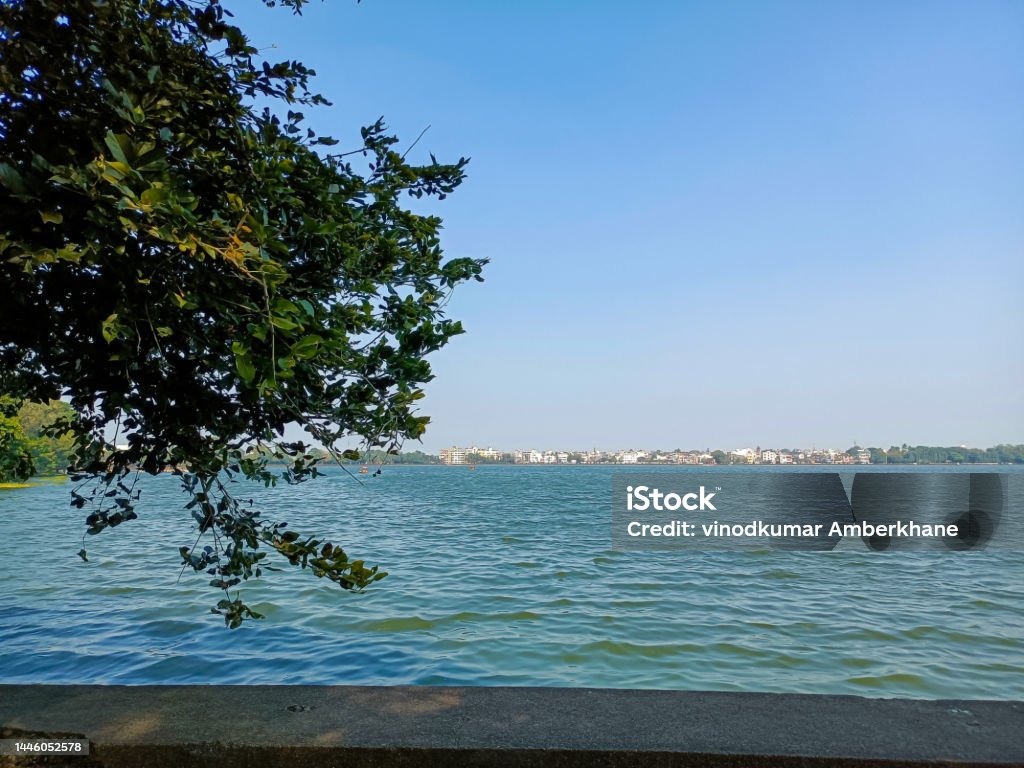 Stock photo of beautiful Rankala lake, old stone retaining wall around the lake, branches of the green tree overhanging on water . Picture captured under bright sunny light at kolhapur, Maharashtra. Atmosphere Stock Photo