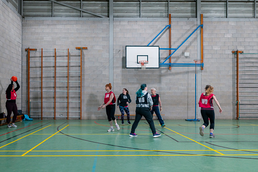 A wide shot of a group of women wearing sports clothing and netball bibs in a sports hall in Newcastle Upon Tyne. One woman takes a throw-in as they are playing a game of netball.