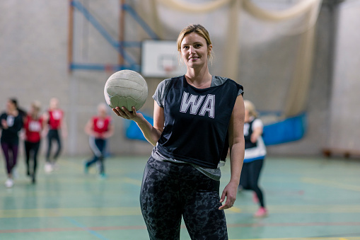 A portrait of a woman wearing sports clothing in a sports hall in Newcastle Upon Tyne. She looks and smiles at the camera as she holds a netball and wears a wing-attack sports bib.