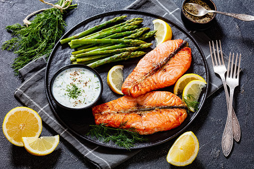 roast salmon steaks with boiled asparagus, fresh lemon and yogurt dill sauce on black plate on concrete table with forkss, landscape view from above