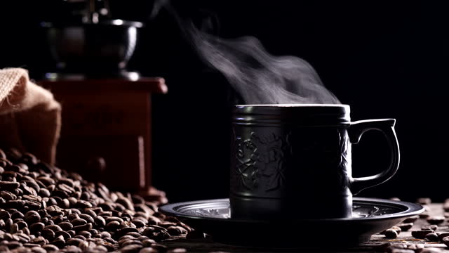 Coffee Cup, Coffee Beans. Close-up beautiful natural steaming smoke from ceramic black coffee mug with vintage grinder espresso on dark background, 4K slow motion. Hot Drink Concept