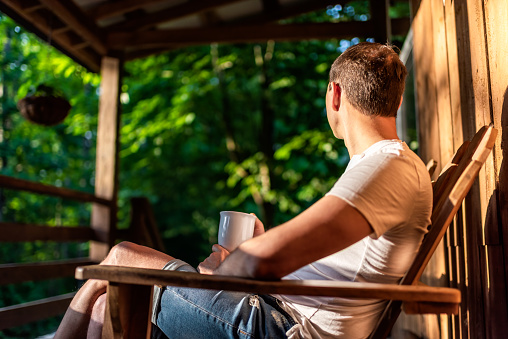 Man sitting on rocking chair on porch of rustic countryside home house wooden cabin cottage, watching sunrise in morning drinking coffee from cup mug