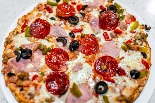 Macro closeup of cooked baked pepperoni black olives ham, mozzarella cheese pizza pie on white plate with tomato sauce and green red bell peppers