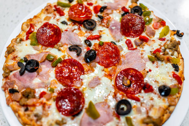 macro closeup of cooked baked pepperoni black olives ham, mozzarella cheese pizza pie on white plate with tomato sauce and green red bell peppers - pepperoni pizza green olive italian cuisine tomato sauce imagens e fotografias de stock