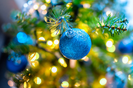 Macro closeup of colorful blue decorative ornament hanging on New Year Christmas tree with light illuminations in winter with bokeh circles in background