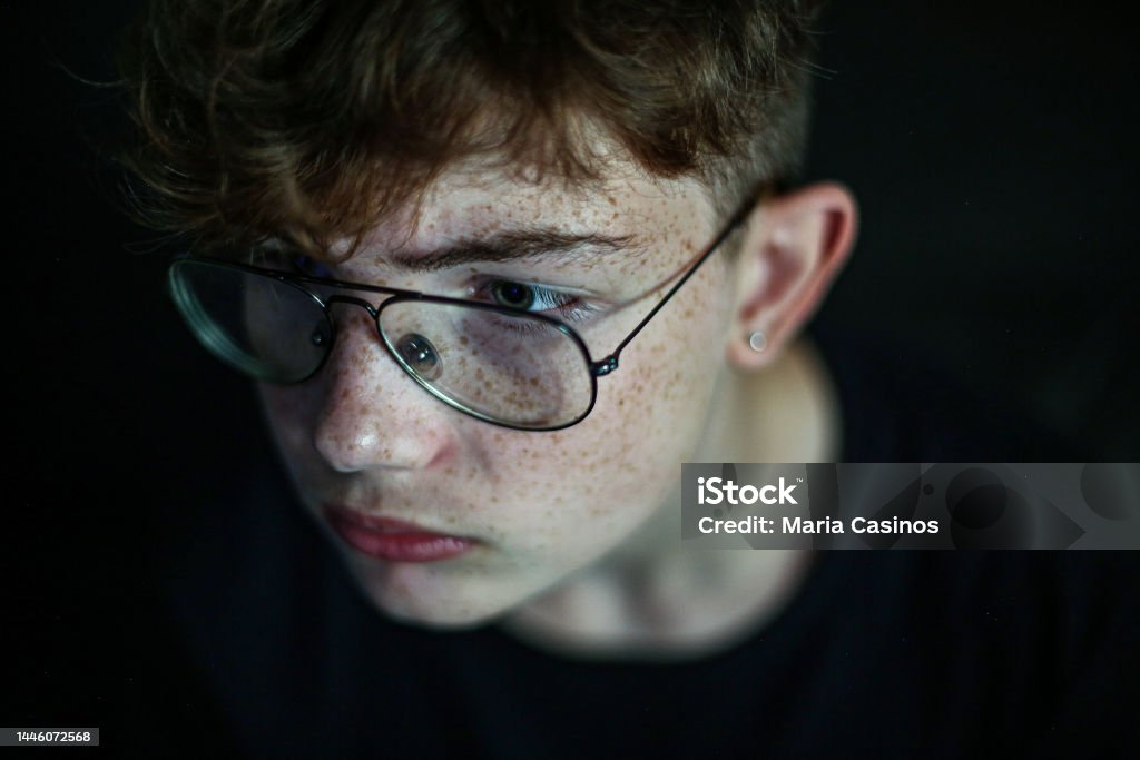 Close-up of  teenager boy, redhead with freckles and glasses, on black background Close-up of red-haired teenager with freckles and glasses, very serious looking probably at a screen, on black background Black Background Stock Photo