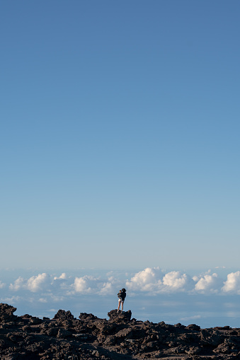 Wide view, rear shot of an active multiracial mother holding her toddler daughter on her hip while admiring the view from above the clouds at the top of Mount Haleakala on Maui, Hawaii. The adventurous family is hiking while on vacation.
