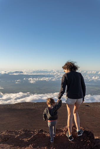 Medium range, rear shot of an active multiracial mother holding her toddler daughter's hand while admiring the view from above the clouds at the top of Mount Haleakala on Maui, Hawaii. The adventurous family is hiking while on vacation.